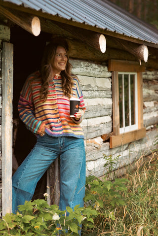 Women wearing a rainbow cardigan and wide leg jeans drinking coffee standing beside a sugar shack.
