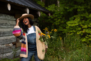 Women carrying a bag of flowers in a rainbow cardigan and overalls in a green forest.