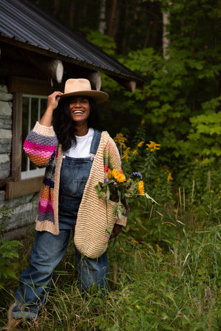 Women carrying a bag of flowers in a rainbow cardigan and overalls in a green forest.