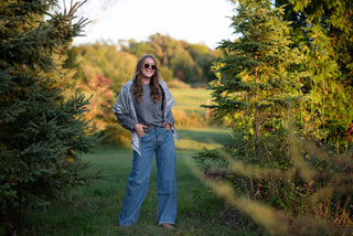 Women wearing a sweater and jeans and a scarf over her shoulders in a grassy patch at a barn. 