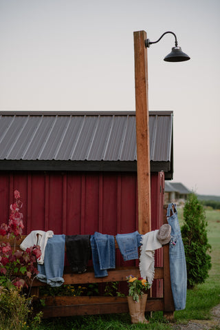 several pairs of jeans folded over and a pair of knee high boots with flowers in them in front of a barn. 