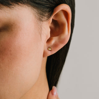 Close-up of an ear wearing a gold heart-shaped earring against a neutral background