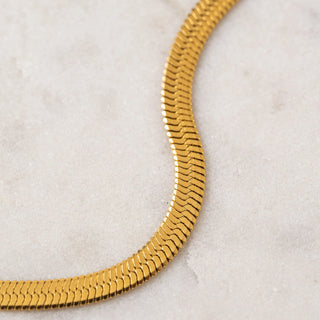 A close-up of a gold-toned snake chain necklace curving across a light beige stone surface.