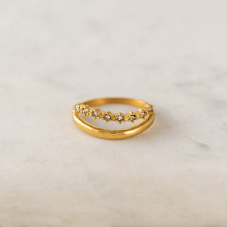 A gold ring with a double-band design and a top band set with small round clear stones, resting on a light beige marble surface.