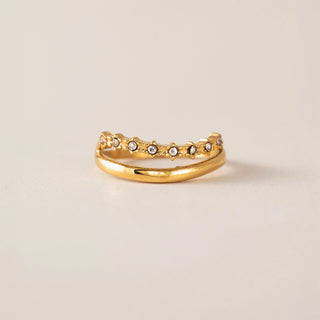 A gold band ring featuring a row of small clear gemstones set in a textured, scalloped border, photographed on a light beige background.