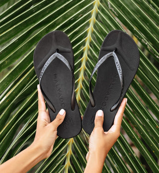 Black sandals held by a person against a green leafy background