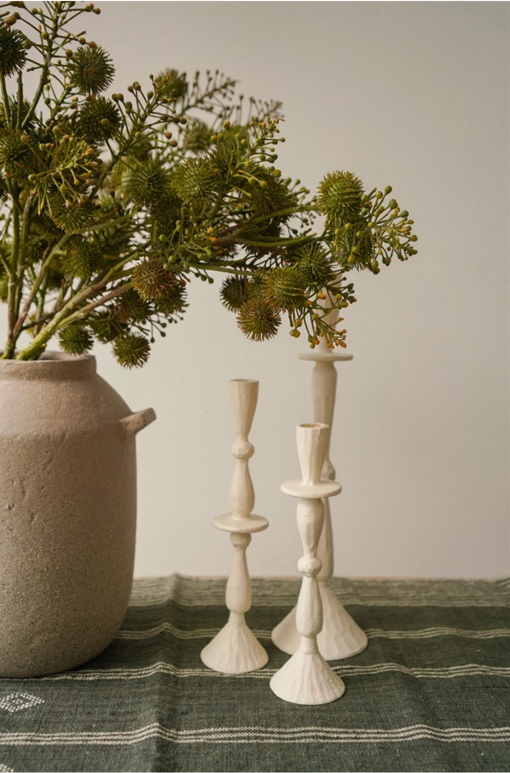 Three white candlesticks on a green textured surface with a vase of greenery in the background.
