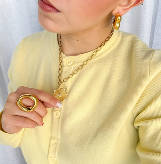 A person wearing A gold pendant necklace with a sunburst design on a white background. 