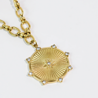 A gold pendant necklace with a sunburst design on a white background. 