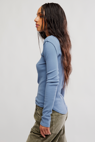 A woman shown in side profile wearing a fitted light blue long-sleeve top and olive green pants against a plain light gray studio background.
