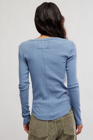 A woman viewed from the back wearing a light blue long-sleeve top with a small rectangular patch detail on the upper back, paired with olive green pants, against a plain light gray studio background.