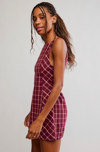 A woman modeling a sleeveless, high-neck mini dress with a burgundy and cream plaid pattern, photographed against a plain light gray studio background.