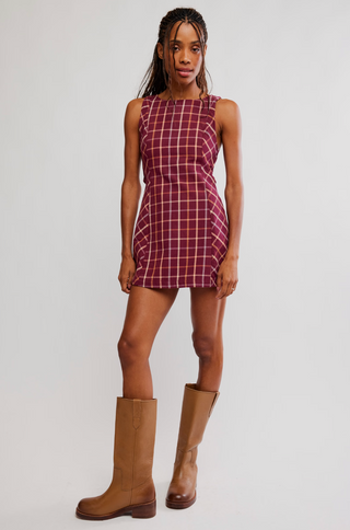 A woman modeling a sleeveless, knee-length plaid dress in burgundy and white, paired with tan knee-high boots, against a plain light gray studio background.