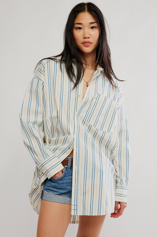 A woman modeling an oversized long-sleeve button-up shirt with vertical blue and green stripes on a cream background, paired with denim shorts, against a plain light gray studio background.