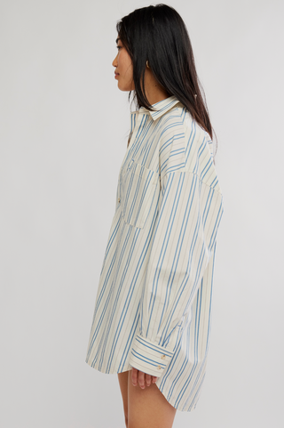 A woman shown in side profile wearing an oversized long-sleeve button-up shirt dress with vertical blue and light gray stripes on a cream background, set against a plain light gray studio backdrop.