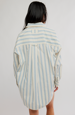 A person shown from the back wearing an oversized striped shirt dress in cream with blue and light green vertical stripes, photographed against a plain light gray studio background.