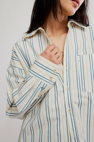 A close-up of a person wearing a cream-colored button-up shirt with vertical blue stripes. The person is shown from the neck down, with one hand adjusting the collar and button area.