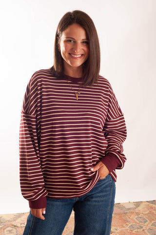 A woman wearing a long-sleeve crewneck top with horizontal burgundy and white stripes, paired with blue jeans, standing against a plain white studio background.