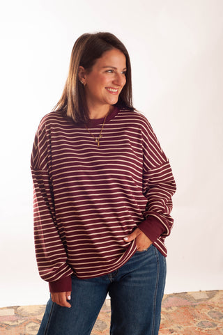 A woman wearing a long-sleeve crewneck shirt with thin horizontal burgundy and white stripes, paired with blue jeans, standing against a plain white studio background.