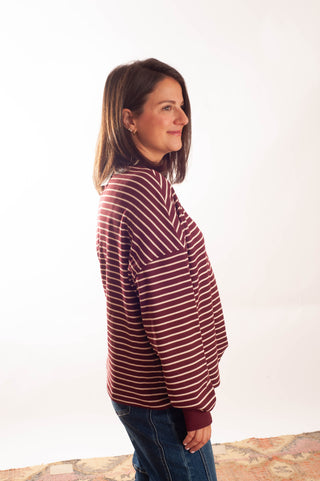 A woman wearing a burgundy and white striped long-sleeve top with blue jeans, standing in profile against a white studio background.