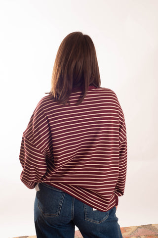 A person viewed from behind wearing a loose-fitting long-sleeve top with thin horizontal burgundy and white stripes, paired with blue jeans, against a plain white studio background.