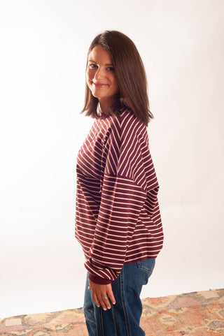 A woman with shoulder-length brown hair is shown from the side, wearing a maroon and white horizontally striped long-sleeve top and blue jeans, standing against a plain white background.