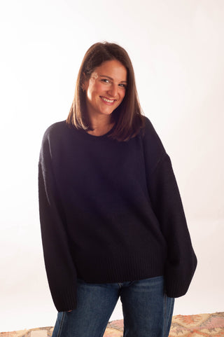 A smiling woman with shoulder-length brown hair wearing an oversized black knit sweater and blue jeans, standing against a plain white studio background.