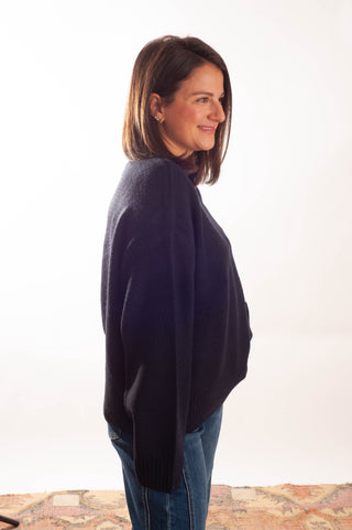 A woman with shoulder-length brown hair is shown in profile wearing a dark navy blue knit sweater and blue jeans against a plain white studio background.