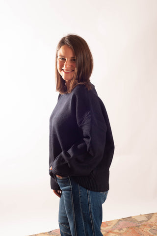 A woman with shoulder-length brown hair wearing a dark navy blue knit sweater and blue jeans, standing against a plain white studio background.