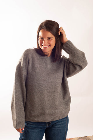A smiling woman with dark brown hair wearing an oversized, heathered gray knit sweater and blue jeans, posing against a plain white studio background.