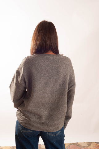 A woman viewed from behind wearing a heather gray knit sweater and blue jeans, standing against a plain white studio background.