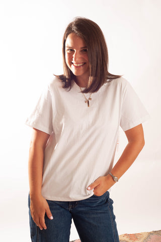 A woman with shoulder-length brown hair smiling while wearing a white short-sleeve crewneck t-shirt and blue jeans, standing against a plain white background.