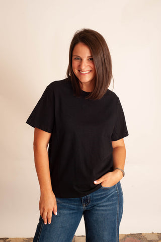 A woman with shoulder-length brown hair smiling while wearing a plain black short-sleeve crewneck t-shirt and blue jeans, standing against a plain off-white wall.