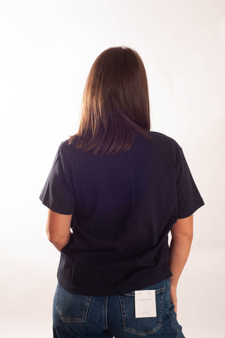 A woman viewed from behind wearing a dark navy blue short-sleeve top and blue jeans, standing against a plain white studio background.
