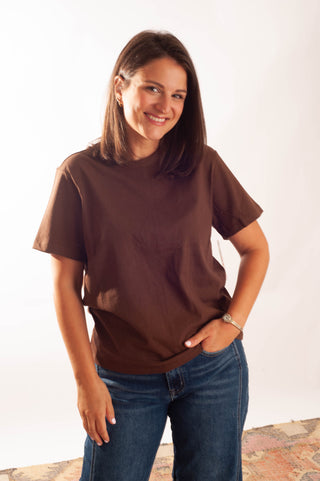 A woman wearing a brown short-sleeve crewneck t-shirt and blue jeans, standing against a plain white studio background.