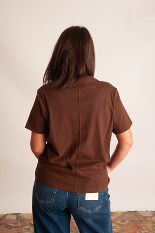 A woman viewed from behind wearing a brown short-sleeve button-up shirt and blue jeans, standing against a plain off-white wall.