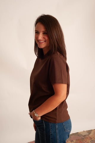 A woman with shoulder-length brown hair is shown in profile, wearing a solid dark brown short-sleeve t-shirt and blue jeans against a plain off-white background.
