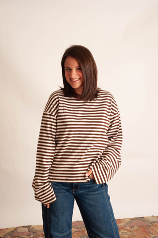 A woman with shoulder-length brown hair smiling while wearing a brown and white horizontally striped long-sleeve top and blue jeans, standing against a plain off-white wall.