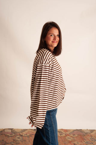 A woman wearing a brown and white horizontally striped long-sleeve top and blue jeans, standing against a plain off-white wall.