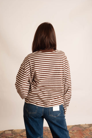 A person viewed from behind wearing a brown and cream striped long-sleeve top and blue jeans, standing against a plain off-white wall.