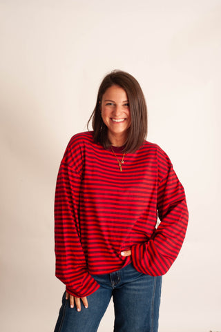 A woman with shoulder-length brown hair smiling while wearing a red and black horizontally striped long-sleeve knit top and blue jeans, standing against a plain off-white studio background.
