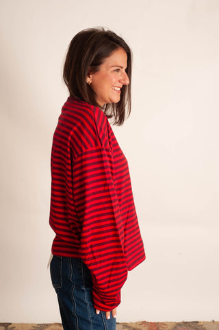 A woman with shoulder-length brown hair is shown in profile, wearing a red and black horizontally striped long-sleeve top and blue jeans against a plain off-white background.