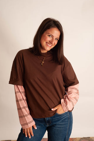 A woman modeling a brown mock neck t-shirt with pink striped long sleeves, paired with blue jeans, against a plain off-white background.