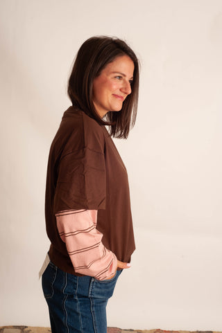 A woman in side profile wearing a brown long-sleeve top with light pink striped elbow-length sleeves and blue jeans, standing against a plain off-white wall.