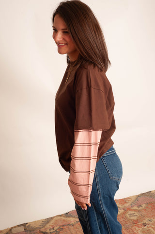 A woman wearing a brown cropped sweatshirt with a contrasting light pink striped long sleeve, paired with blue jeans, standing against a plain off-white wall.
