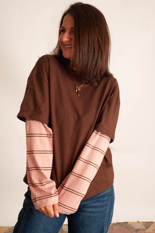 A woman wearing a brown short-sleeve t-shirt layered with a light pink long-sleeve striped shirt, paired with blue jeans, standing against a plain off-white wall.