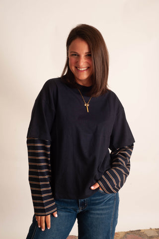 A woman wearing a dark navy long-sleeve top with striped sleeves, paired with blue jeans and a gold key necklace, standing against a plain off-white wall.