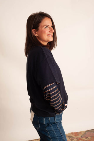 A woman wearing a navy blue sweatshirt with striped sleeves and blue jeans, standing against a plain off-white wall.