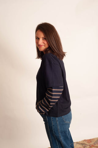 A woman with shoulder-length brown hair is shown from the side, wearing a dark navy blue t-shirt with striped sleeves and blue jeans, standing against a plain off-white background.