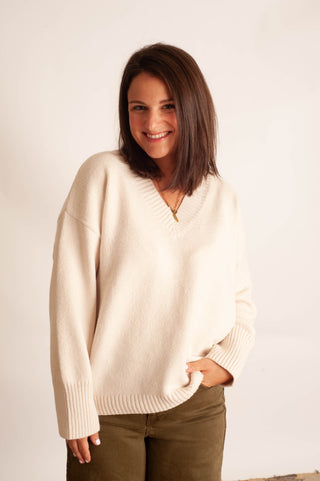 A woman with shoulder-length brown hair smiling while wearing a cream-colored knit V-neck sweater and olive green pants, standing against a plain off-white background.
